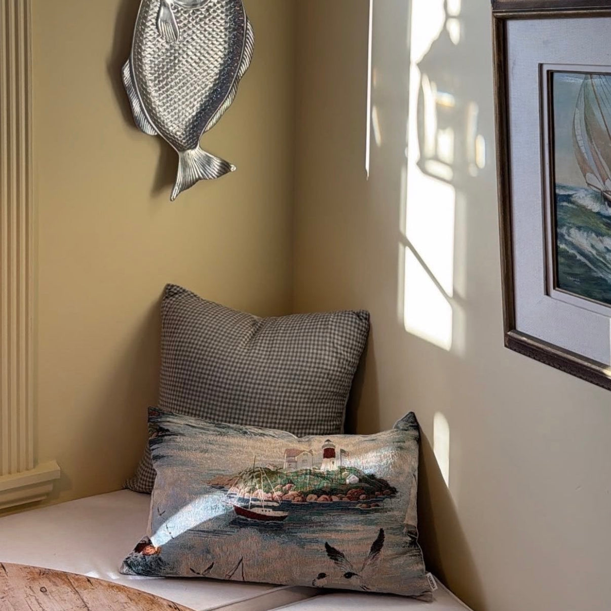 A styled interior corner with a beige wall, a wall-mounted glass shade sconce, a silver fish wall decor piece, and a built-in bench with two pillows, including one with a nautical scene print.